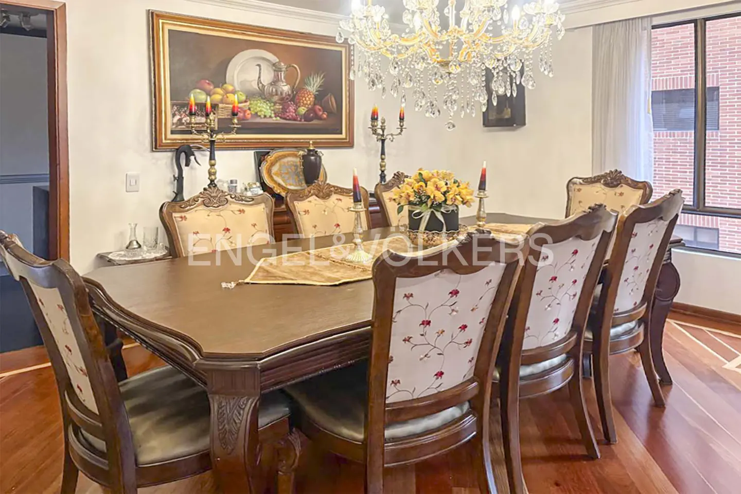 Formal dining room with a long wooden table, ornate chairs, chandelier, and fruit painting.
