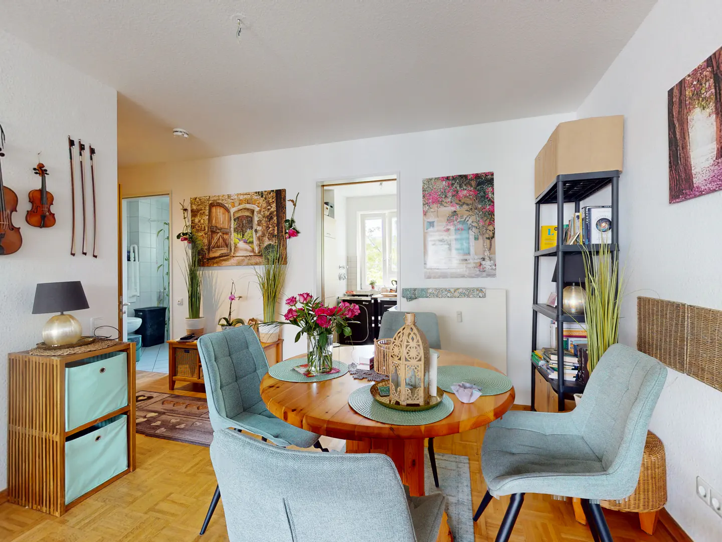 A bright dining area with a round wooden table, four blue chairs, and floral decorations. Artwork adorns the white walls, and a bookshelf stands nearby.