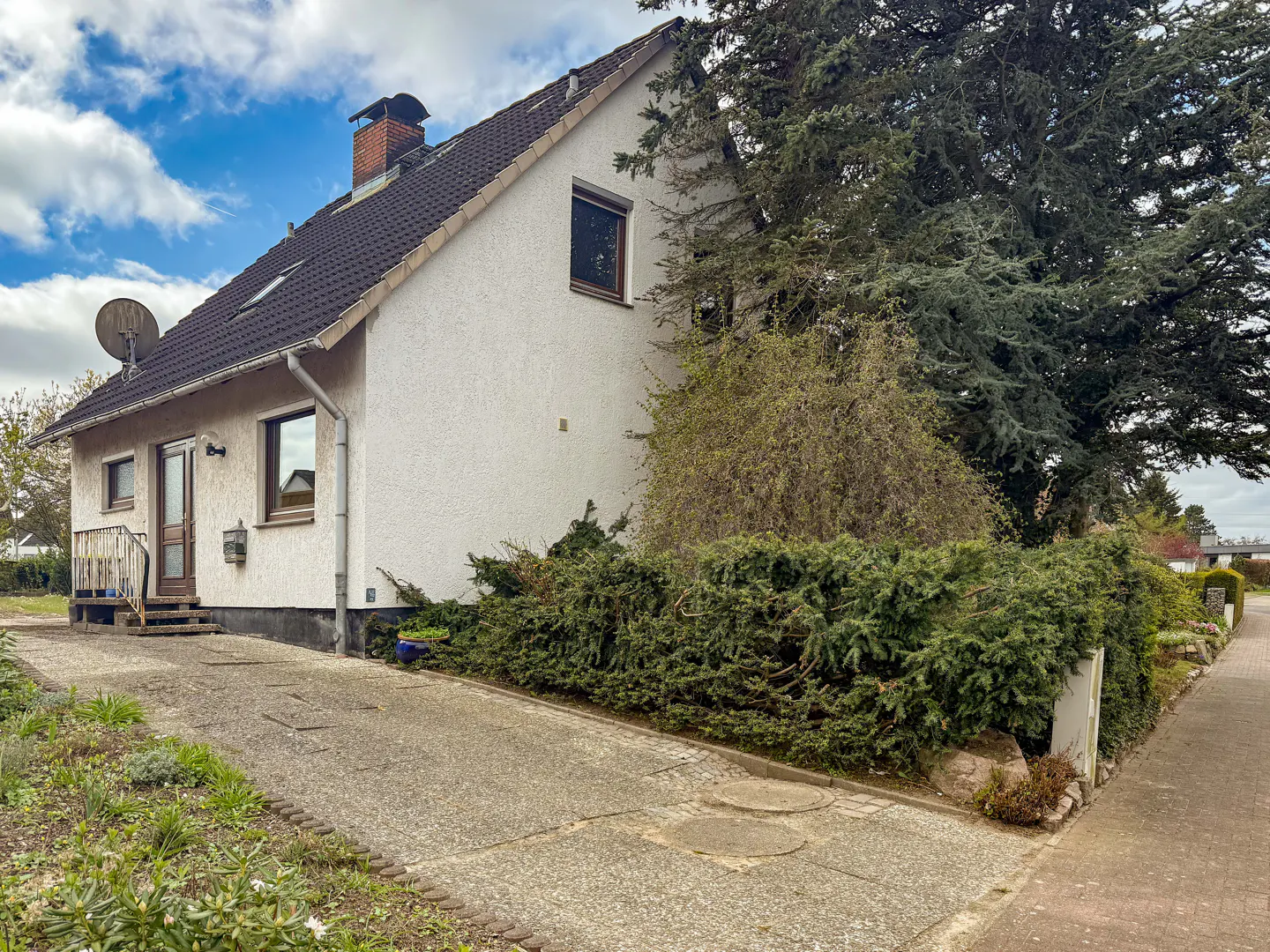 A white two-story house with a dark tiled roof, a brick chimney, and a concrete driveway next to a large evergreen tree.