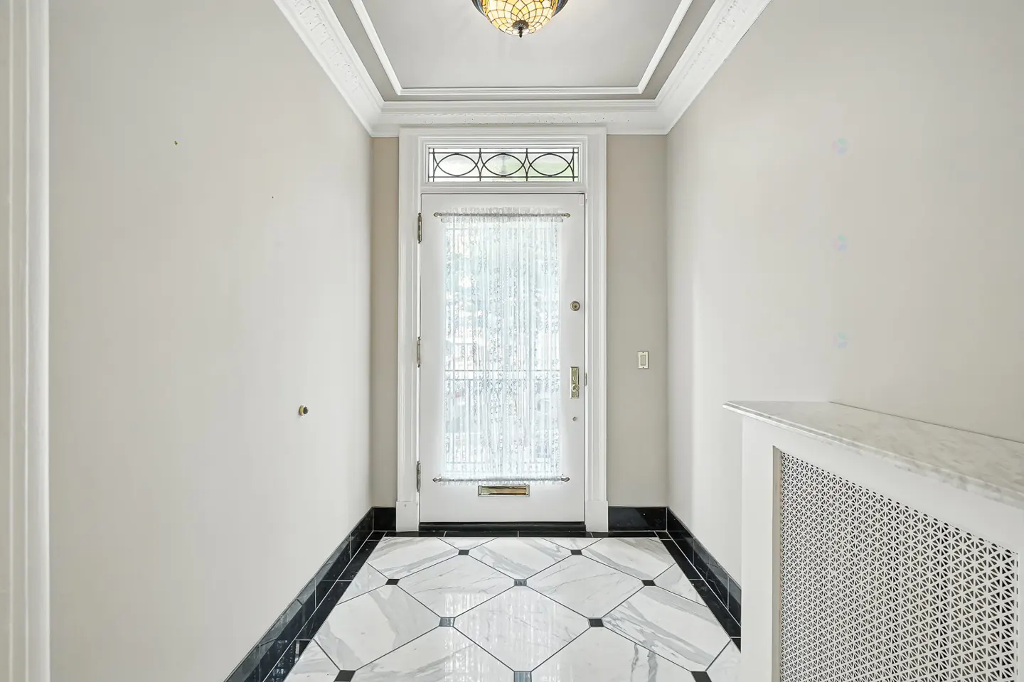 A bright foyer with white walls, a white front door with sheer curtains, and a black and white tiled floor.