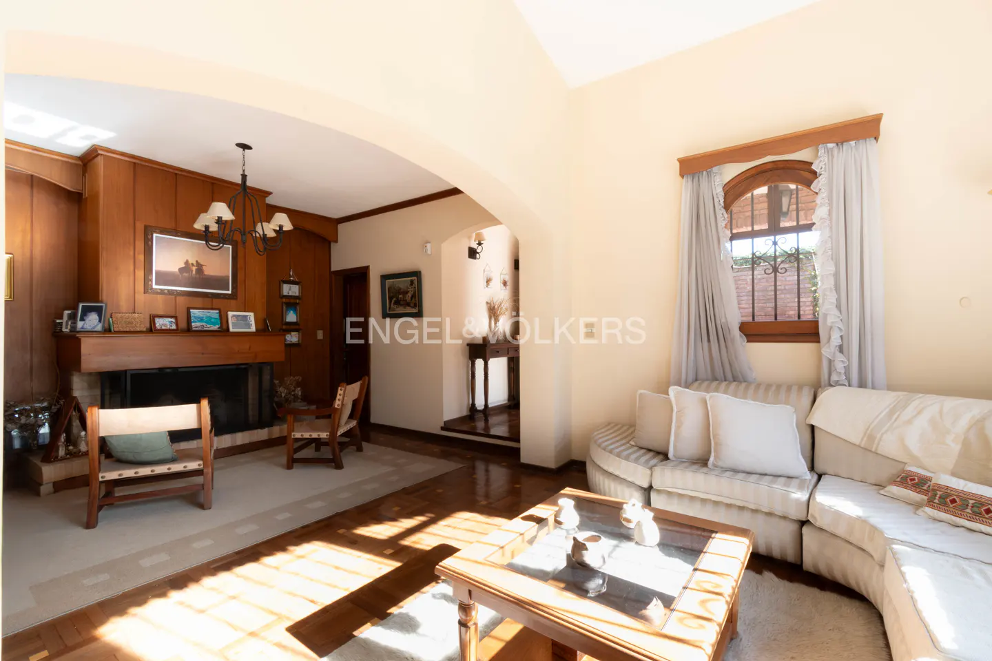 Living room with wood paneling, fireplace, and beige sectional sofa. Sunlight streams through a window with sheer curtains.
