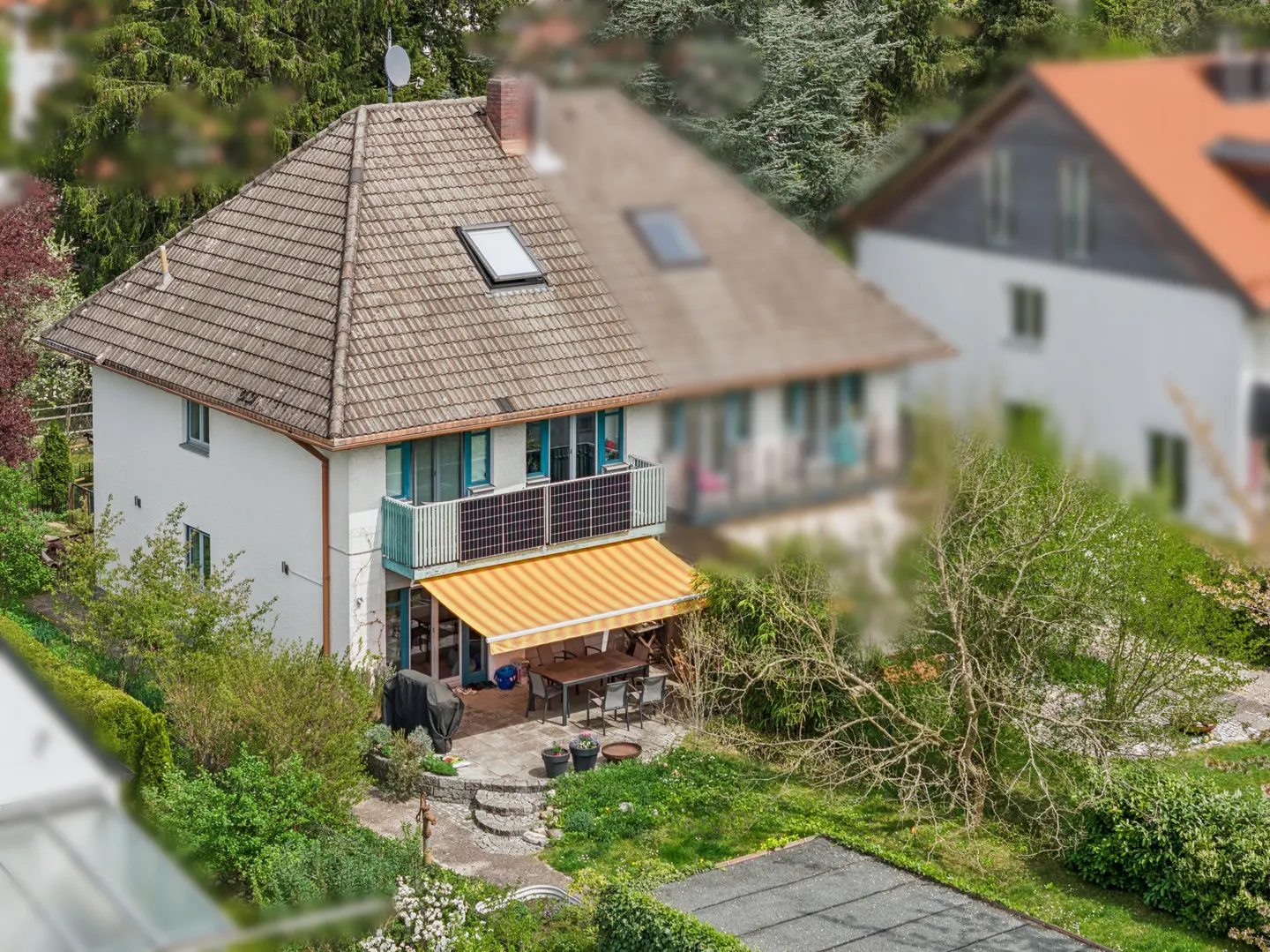 Two-story white house with a brown tile roof, skylight, and solar panels on the balcony. A yellow awning covers a patio with a table and chairs. Green foliage surrounds the house.