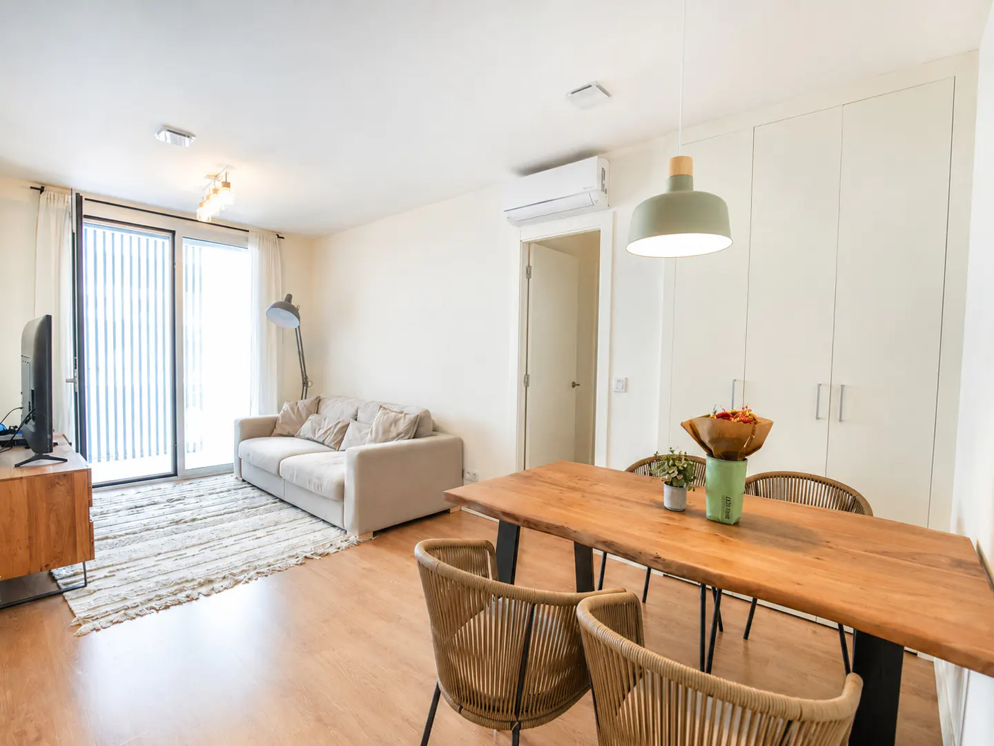 Bright living and dining area with a beige sofa, wooden table with woven chairs, and a large window.