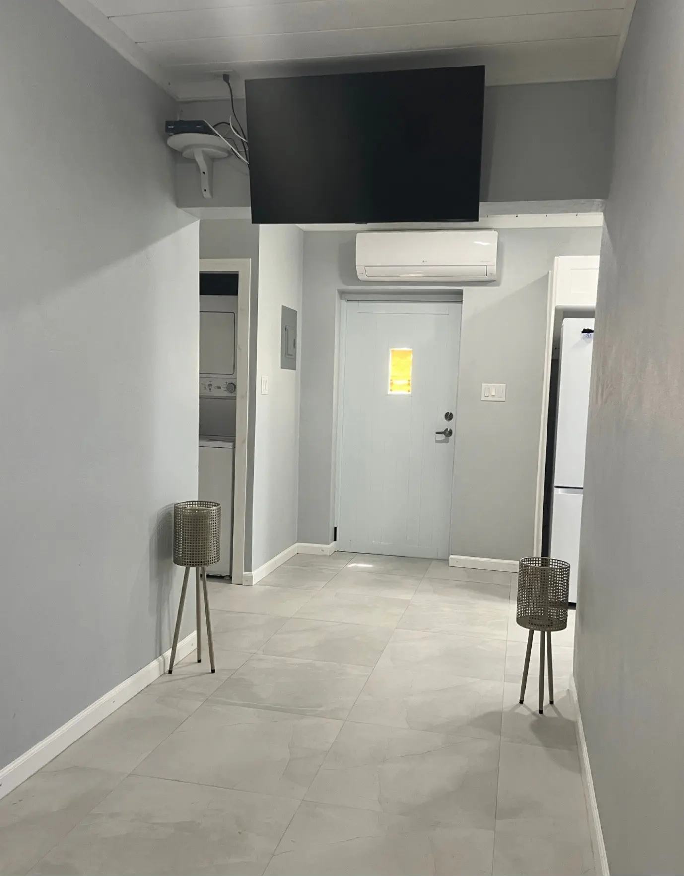 A gray room with a TV, AC unit, and a door with a yellow square. There are two plant stands, a washer/dryer, and a refrigerator.