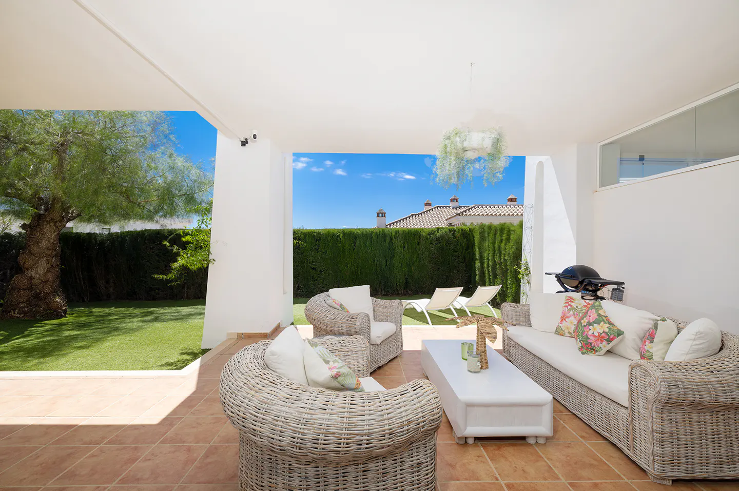 Covered patio with wicker furniture, white cushions, and a white coffee table on terracotta tile. A grill sits near a white wall. Green lawn and blue sky visible.