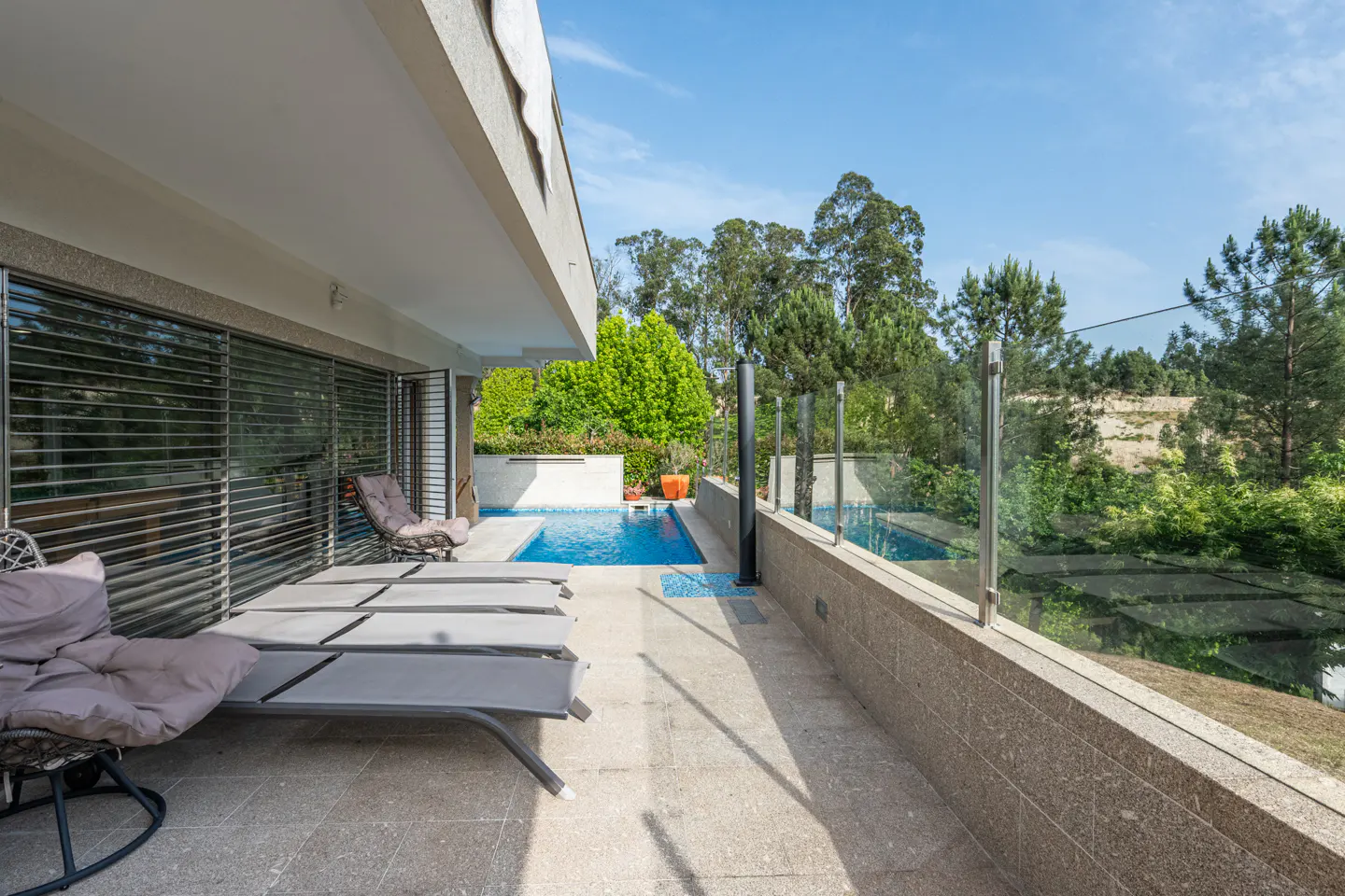Patio with lounge chairs overlooking a blue swimming pool and green trees under a blue sky.