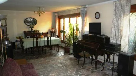 A living room with a dining table, piano, TV, and brick floor. A chandelier hangs above the dining table. Curtains frame the windows.