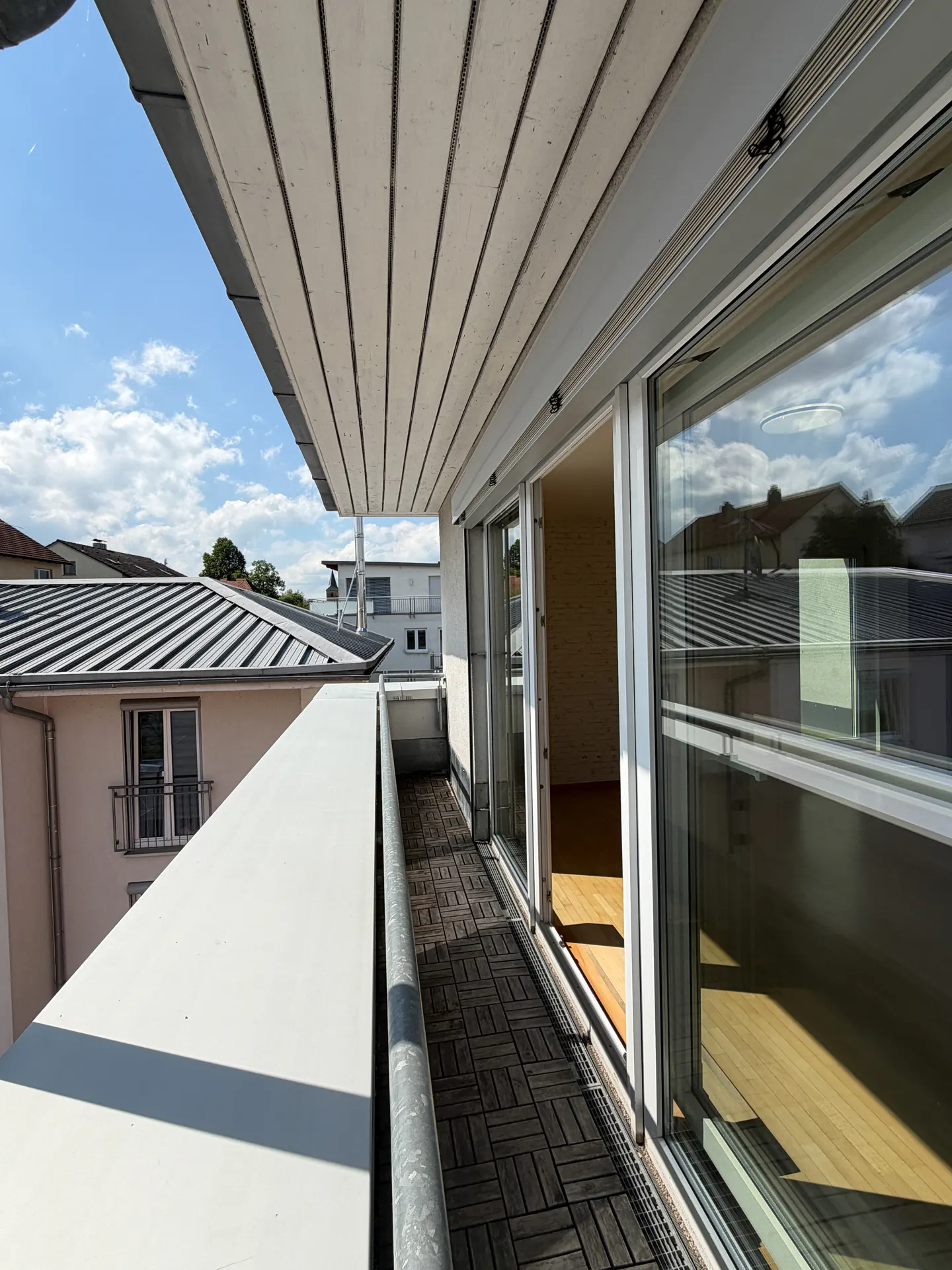Balcony view with wood-look tile floor, white railing, and sliding glass doors. Exterior view of rooftops and blue sky.