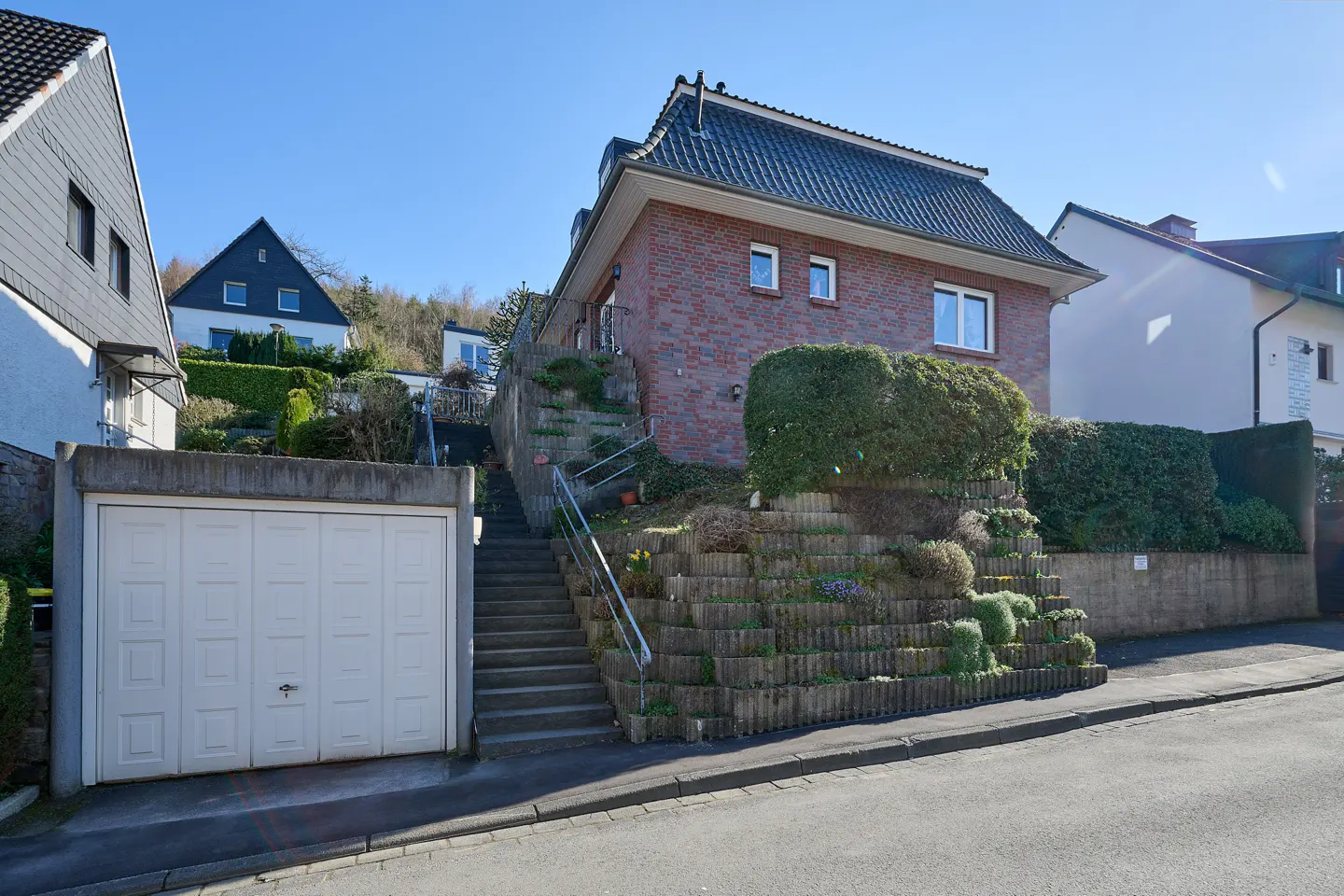 A red brick house with a black roof sits on a hill with a long staircase and a white garage.