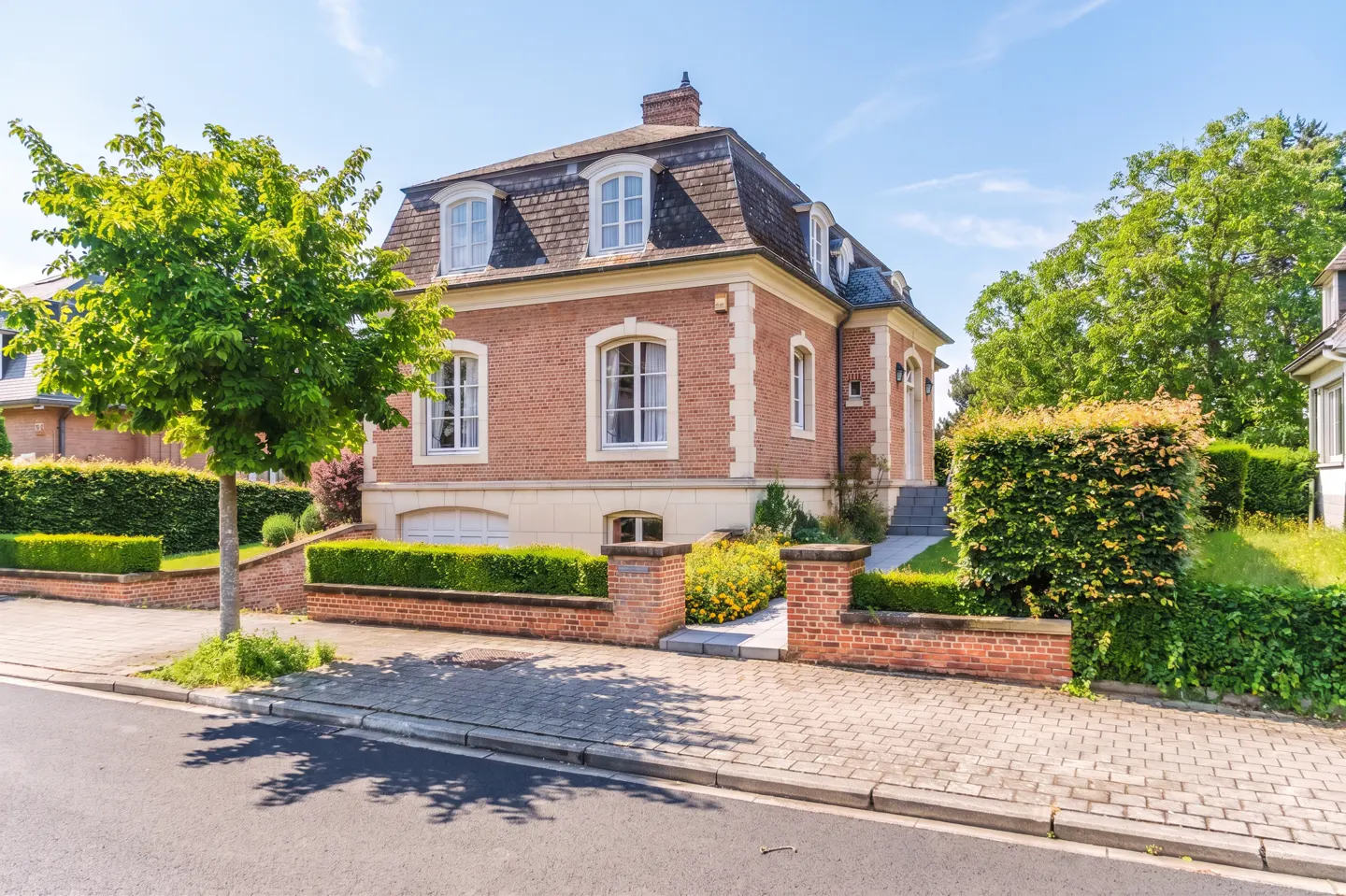 A two-story red brick house with a mansard roof, white trim, and a manicured garden behind a low brick wall.