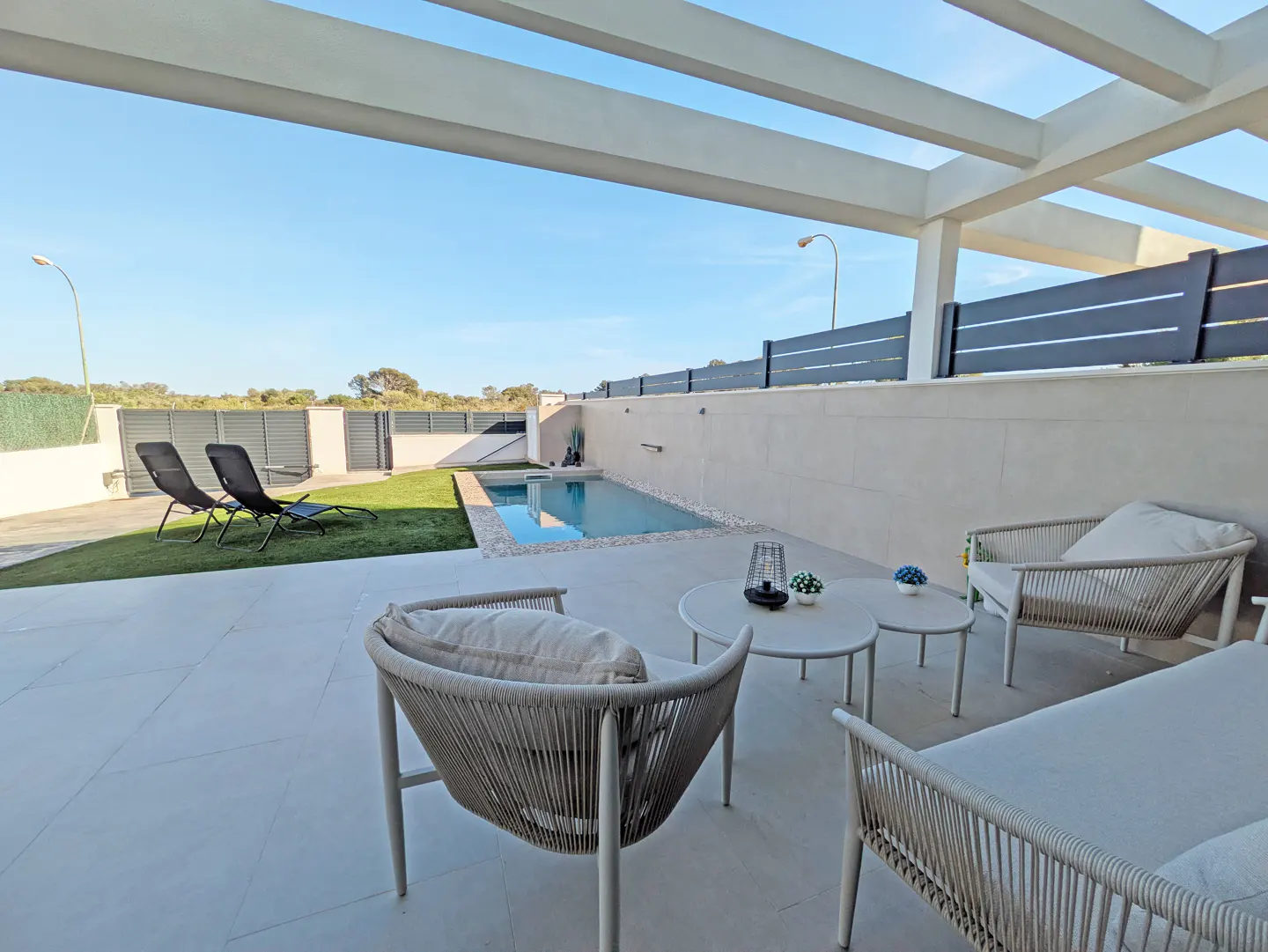 A modern patio with white furniture, a pool, and two black lounge chairs on the grass. The sky is blue and the sun is shining.