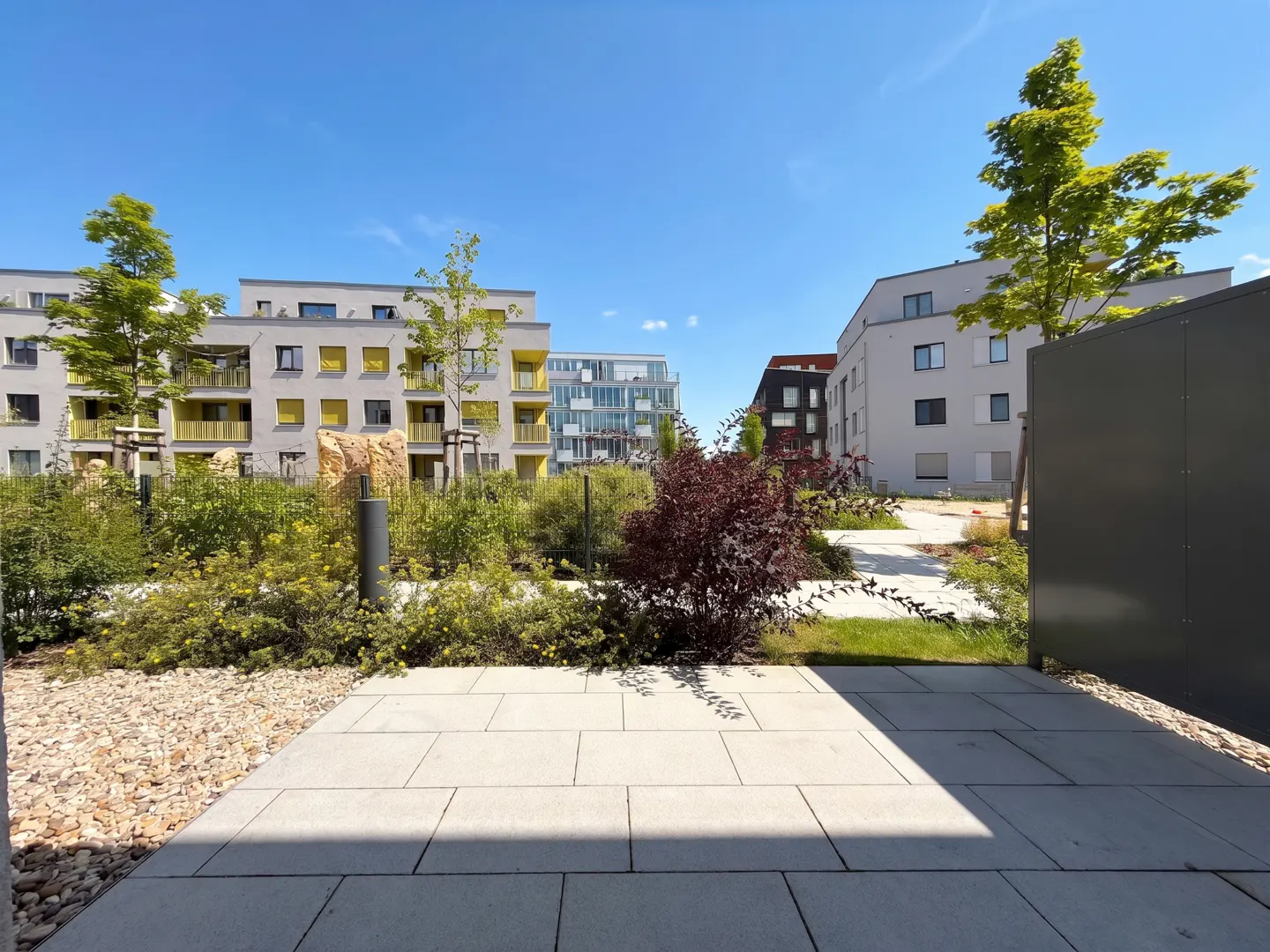 View of modern apartments with yellow balconies, green trees, and a stone pathway under a bright blue sky.