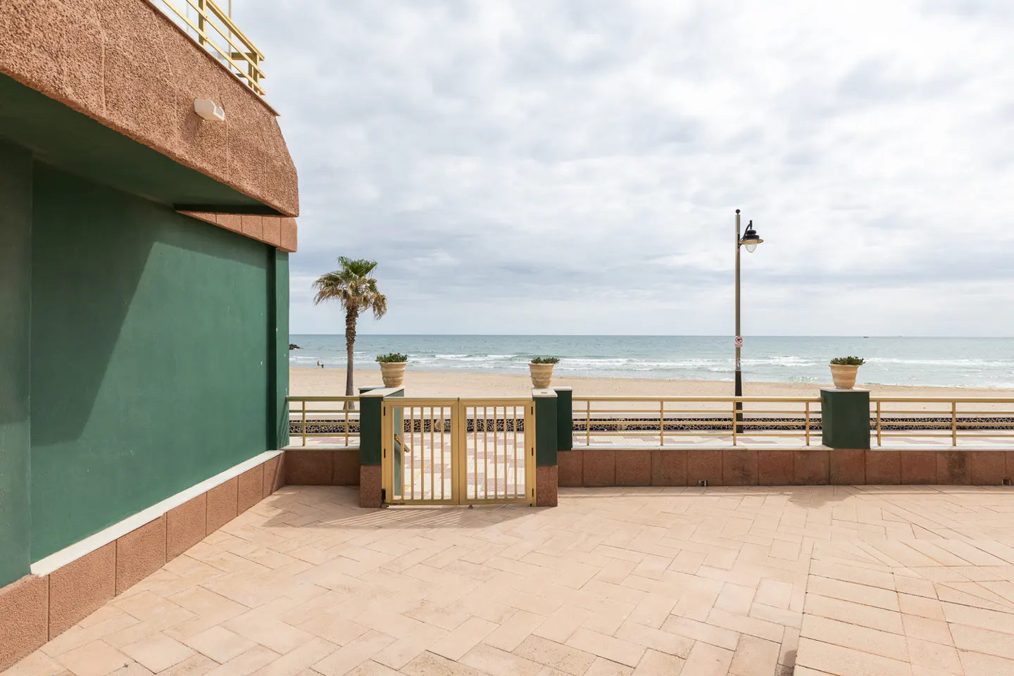 View from a tiled patio with a gold gate leading to a sandy beach and ocean under a cloudy sky. A palm tree and lamppost are visible.