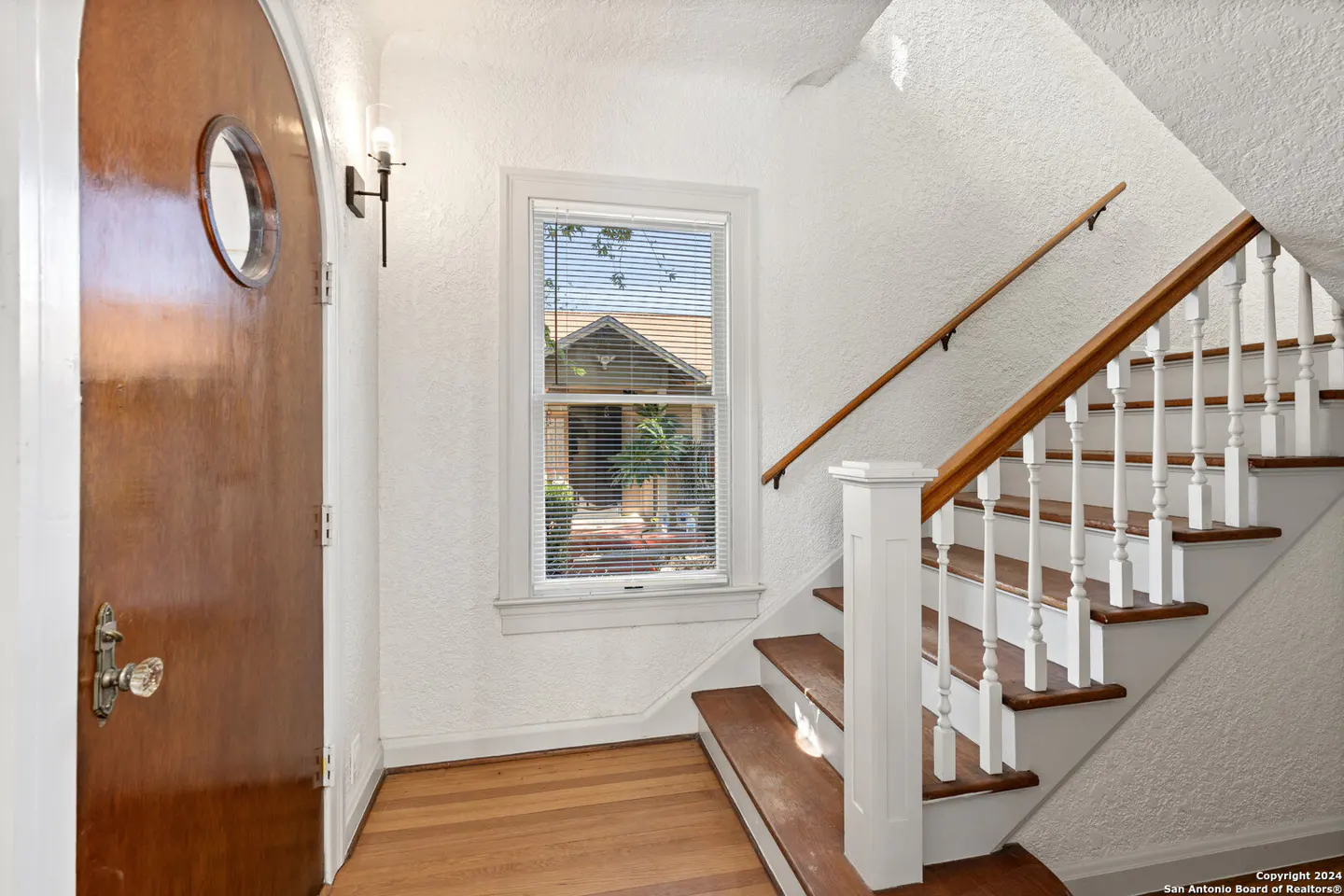 Entryway with a wooden arched door, a window with blinds, and a staircase with white railings and wood steps.