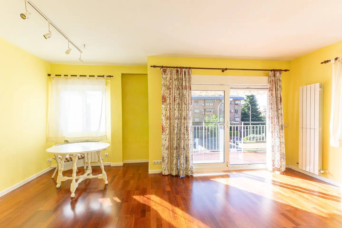 Bright, empty room with yellow walls, wood floors, and a white table. Balcony doors with floral curtains lead to an outdoor view.