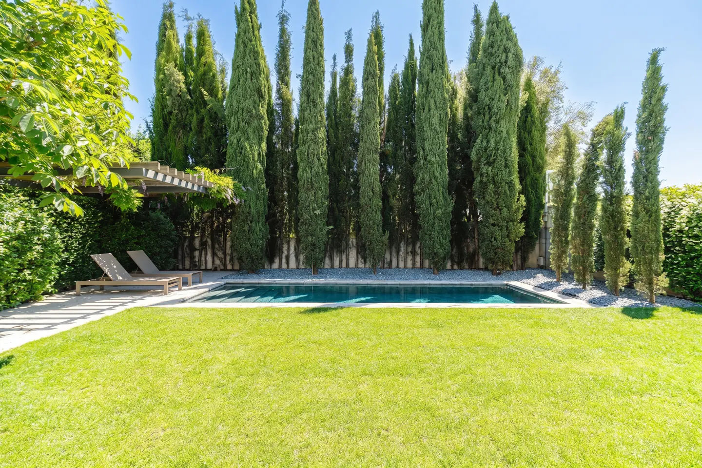 A backyard with a pool, green grass, tall trees, and two lounge chairs.