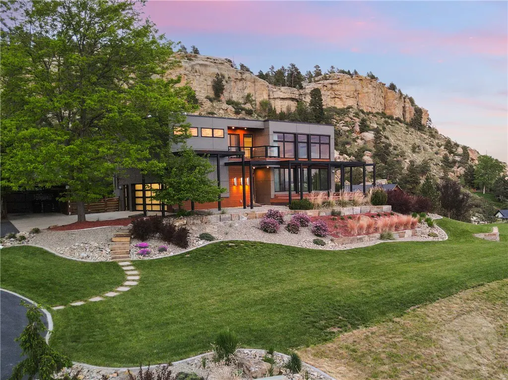 Modern two-story home with a black metal balcony, large windows, and a green lawn, set against a rocky hillside at dusk.