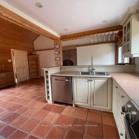 A kitchen with terracotta tile flooring, cream cabinets, stainless steel appliances, and wood beam accents. Wine glasses hang above the sink.