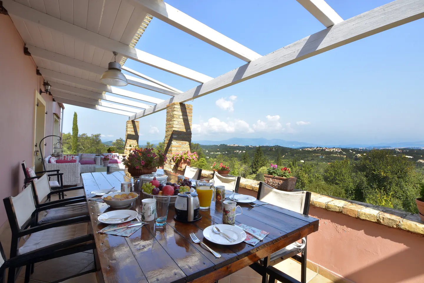 Outdoor dining area with a wooden table set for breakfast, under a white pergola with a scenic view of green hills and blue sky.
