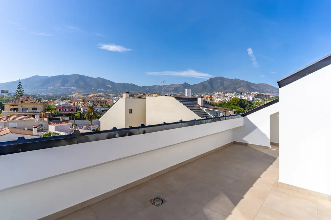 Spacious rooftop terrace with white walls and tiled floor, overlooking a town with mountains under a clear blue sky.