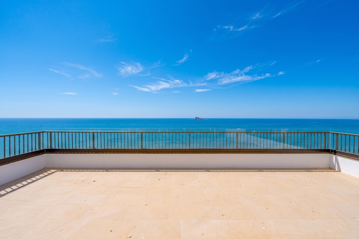 Spacious balcony with beige tile flooring and a metal railing overlooking the blue ocean under a clear sunny sky.