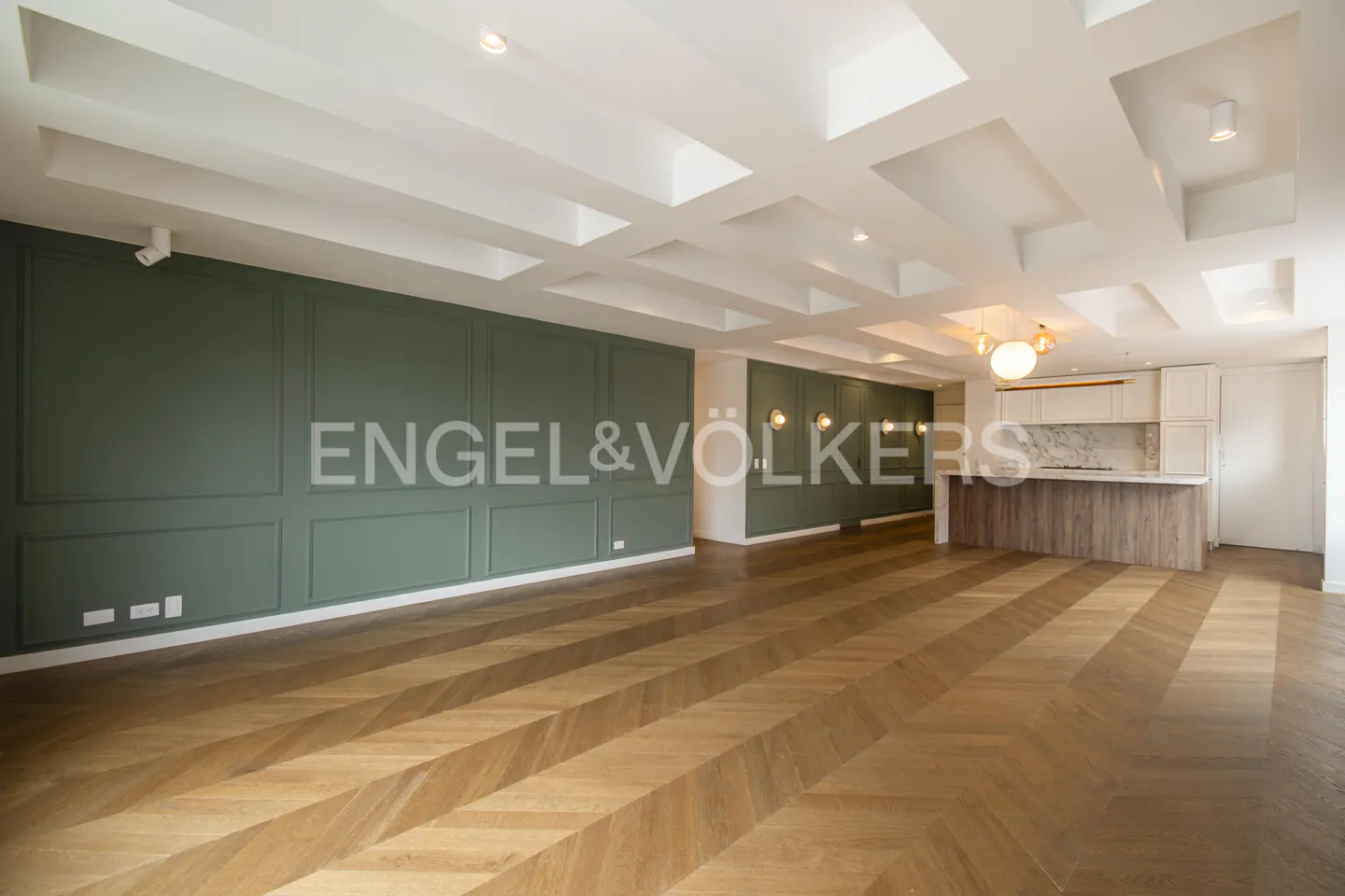 An open-concept living space with herringbone wood floors, green paneled walls, and a white coffered ceiling. A kitchen island is visible in the background.