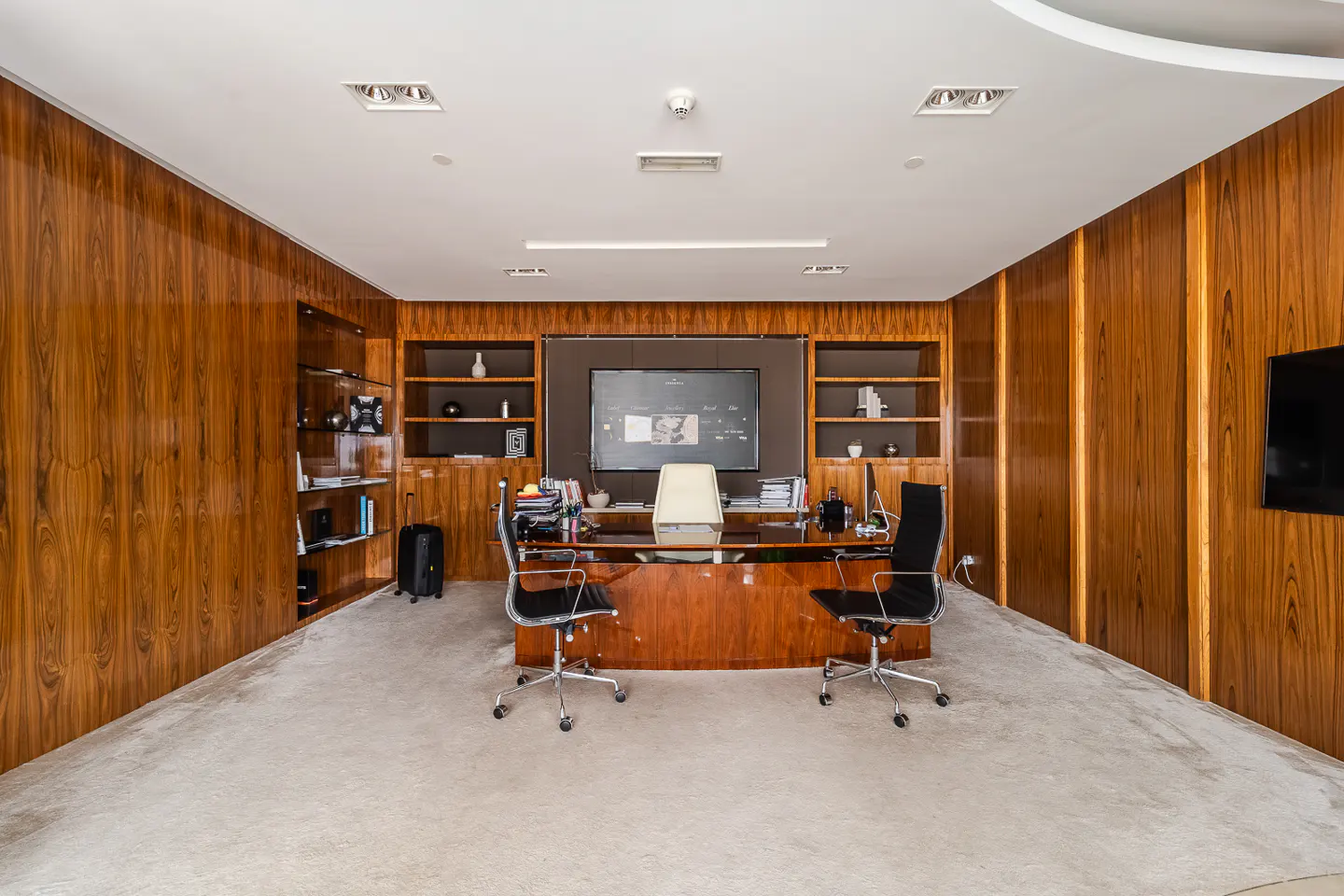 A wide shot of a wood-paneled office with a large desk, two black chairs, and a beige carpet. A TV screen is mounted on the wall above the desk.