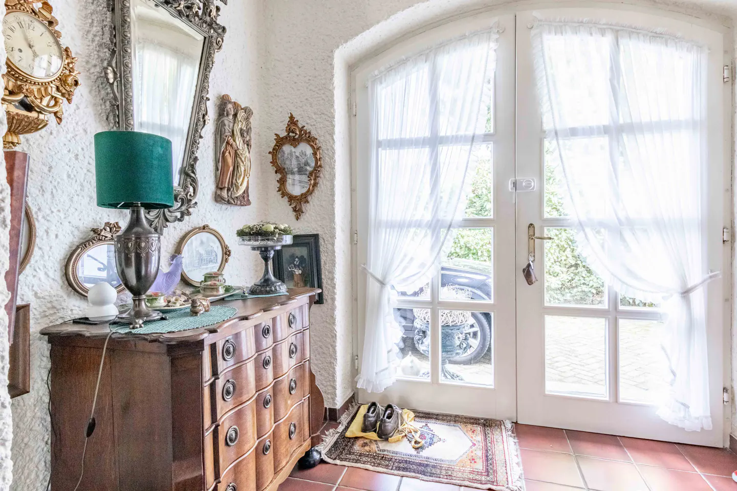 Foyer with a wooden dresser, green lamp, and ornate mirror. White double doors with sheer curtains lead outside to a car.
