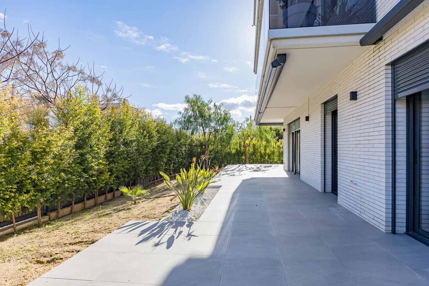 Modern home exterior with a gray tiled patio, white brick, and green hedges under a sunny sky.