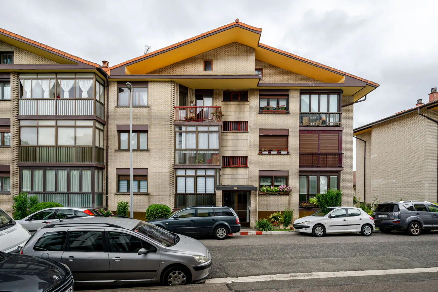 Exterior view of a multi-story brick apartment building with enclosed balconies and parked cars on the street in front.