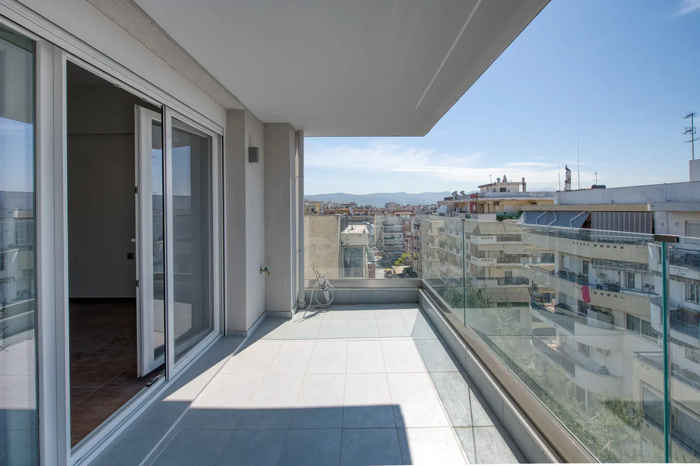 A bright, modern balcony with glass railings overlooks a cityscape under a clear blue sky. Sliding glass doors lead to the interior.