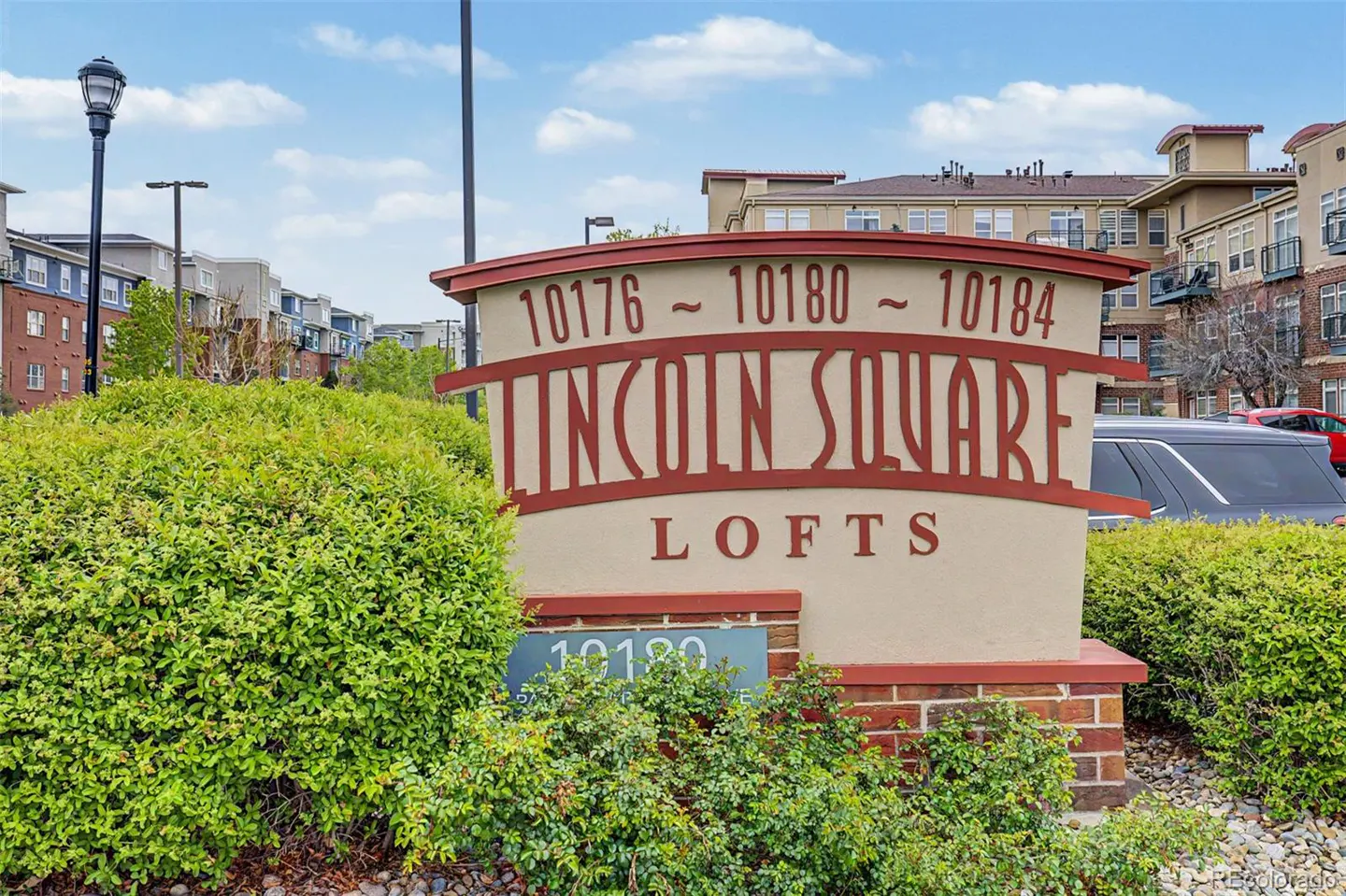 Lincoln Square Lofts sign with address numbers 10176-10184. The sign is beige with red trim and lettering, surrounded by green bushes.