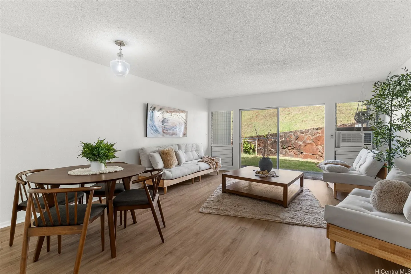 Bright living and dining area with light wood floors, white sofas, a wooden coffee table, and a view of a grassy hill.