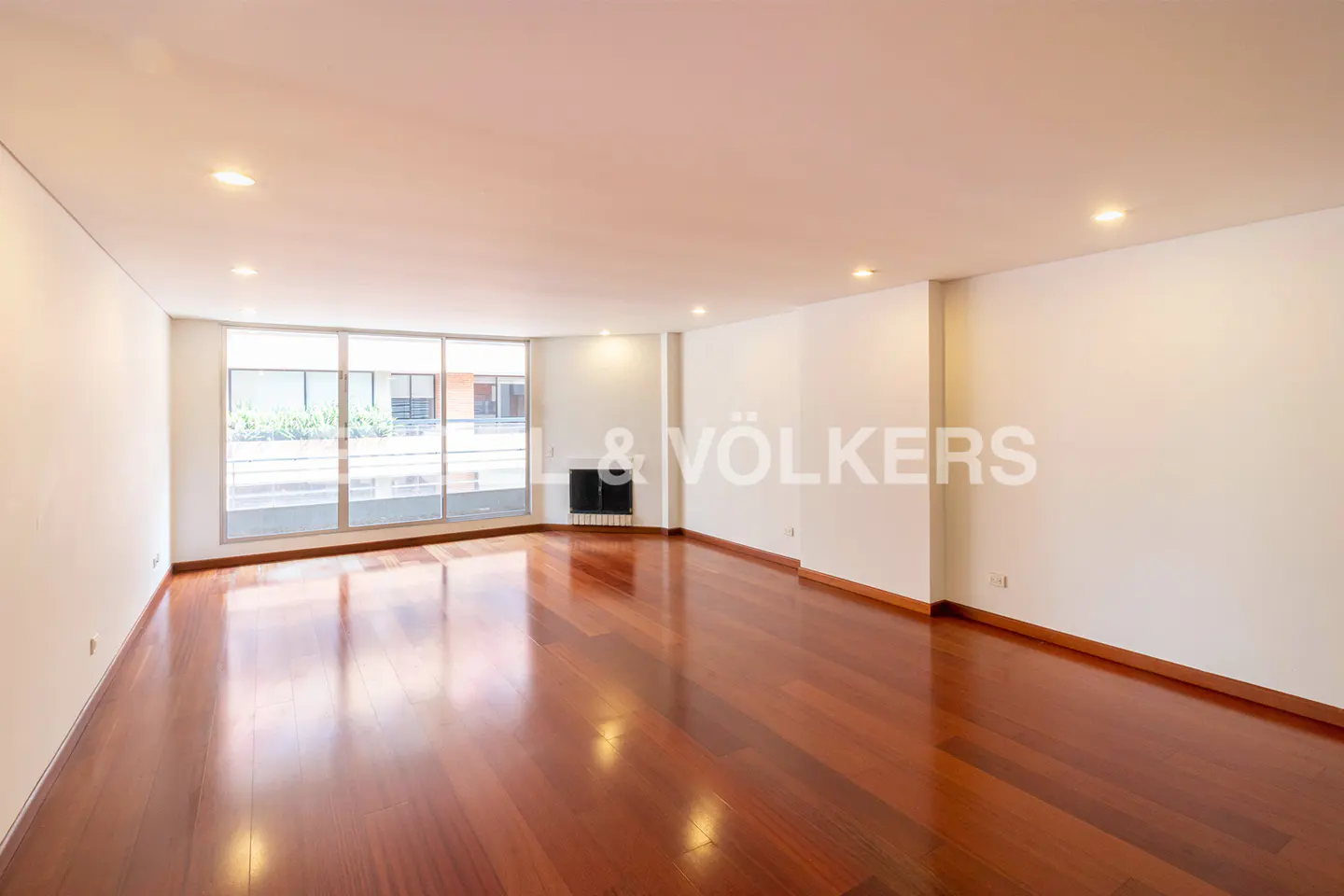 Empty room with hardwood floors, white walls, and large windows. A black fireplace is visible. The Engel & Völkers logo is on the wall.