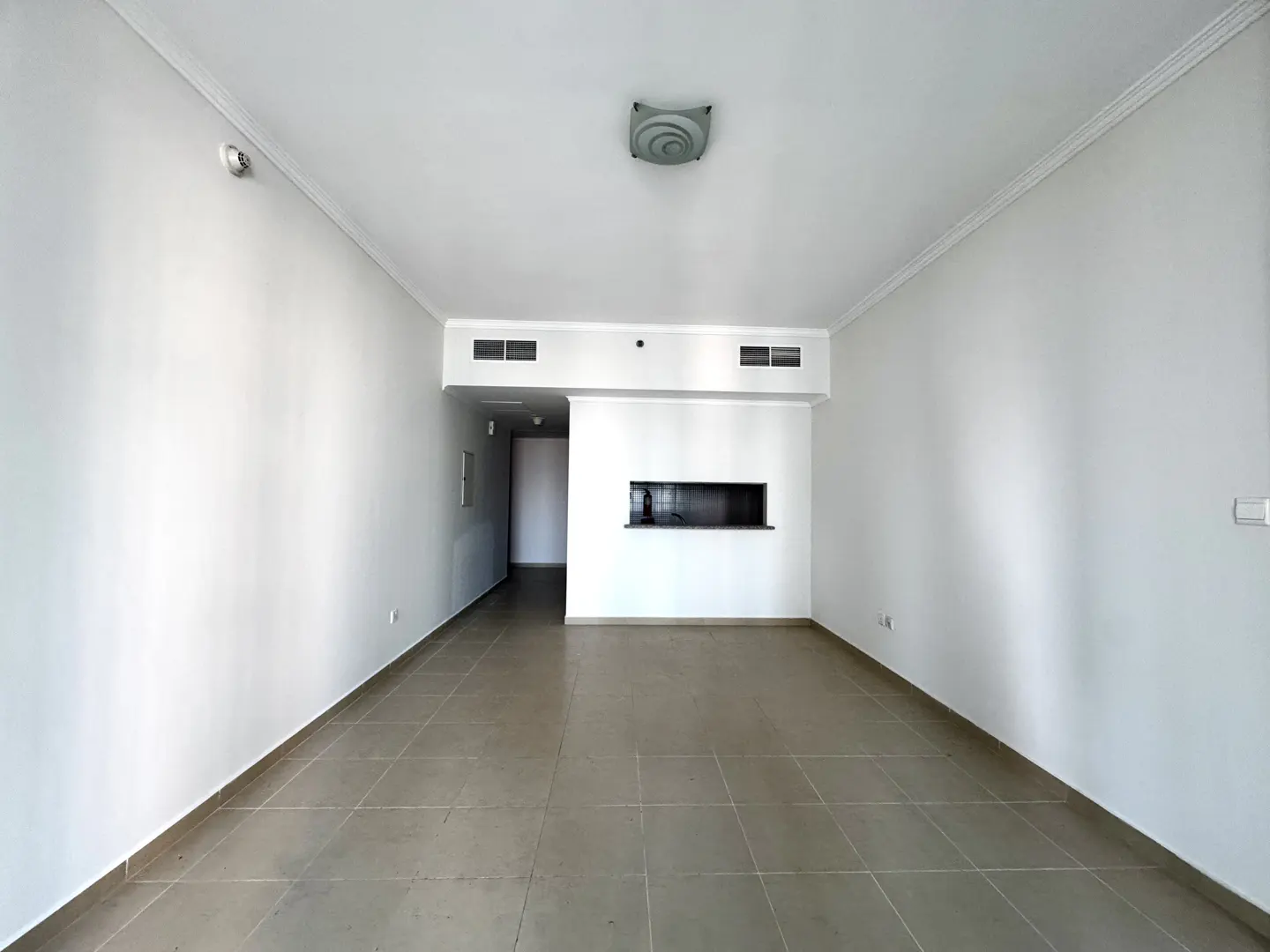 Empty room with white walls, tiled floor, and a kitchen pass-through. A hallway is visible in the background.
