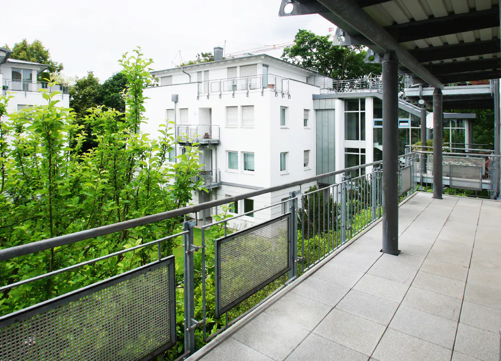 View from a gray tiled balcony with metal railings, overlooking green trees and a white apartment building.