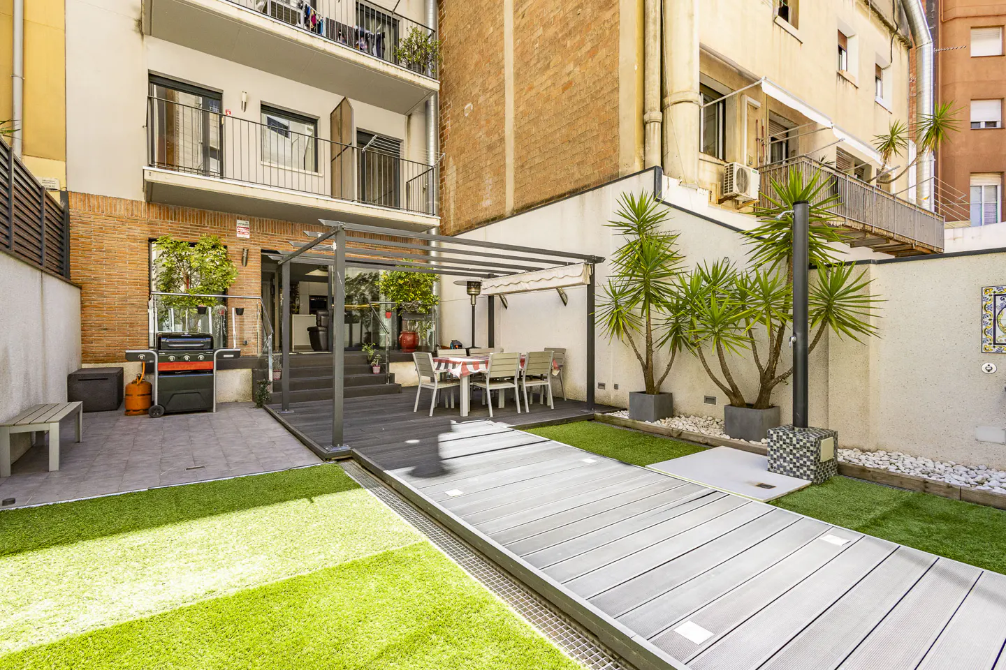 A backyard with a grill, table, chairs, and green grass. The yard is surrounded by buildings.