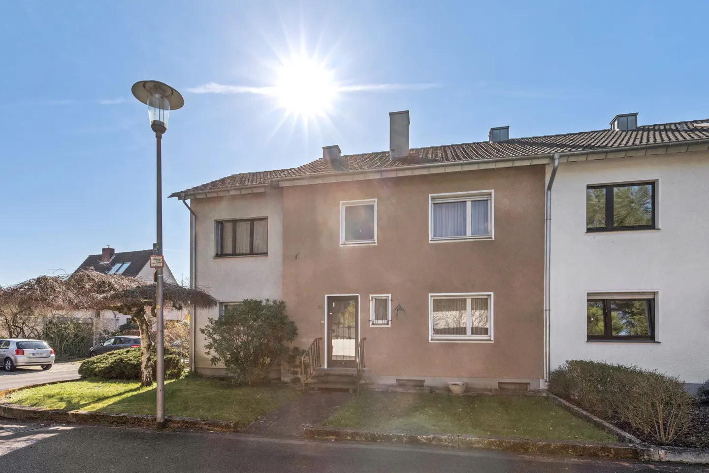 Exterior view of a two-story tan and white townhouse with a front lawn, a street lamp, and a bright sun in a blue sky.