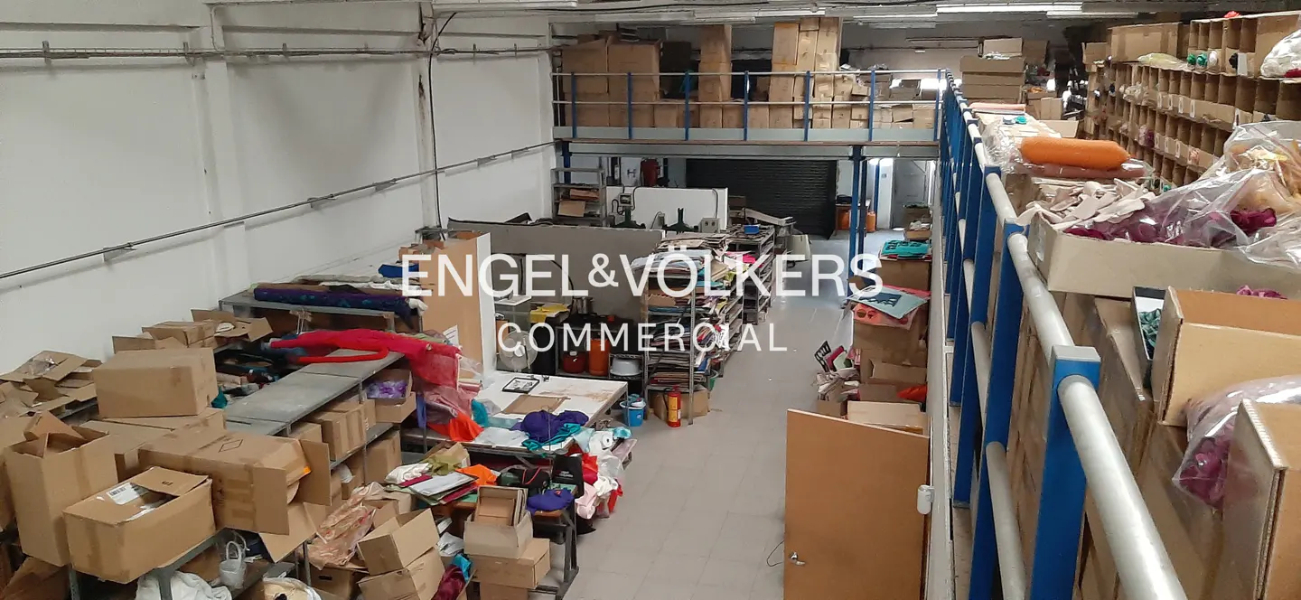 Wide shot of a cluttered warehouse interior with boxes, shelves, and a mezzanine level. The Engel & Volkers Commercial logo is superimposed.