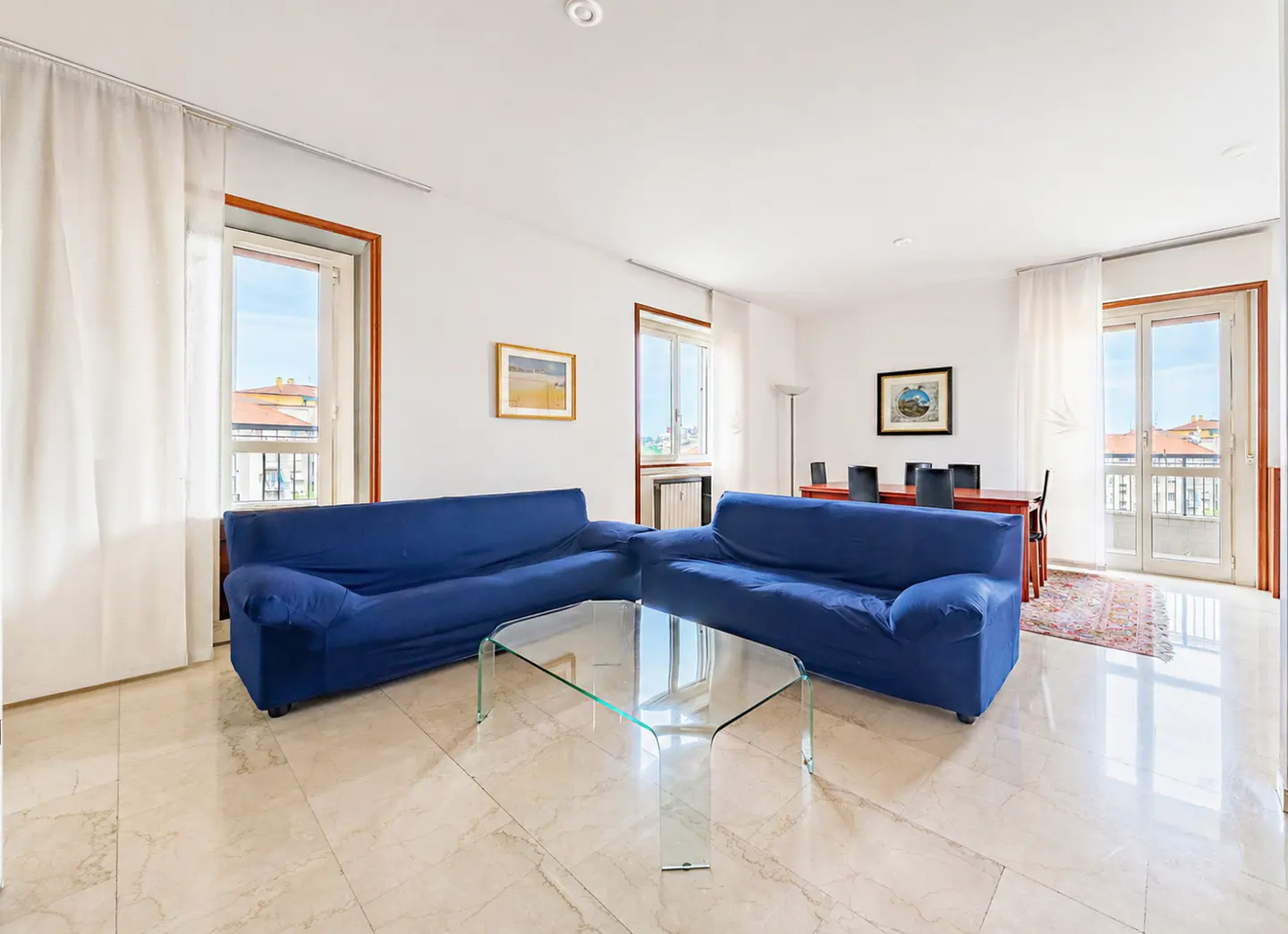 Bright living room with marble floors, two blue sofas, and a glass coffee table. Dining table and chairs visible in the background.