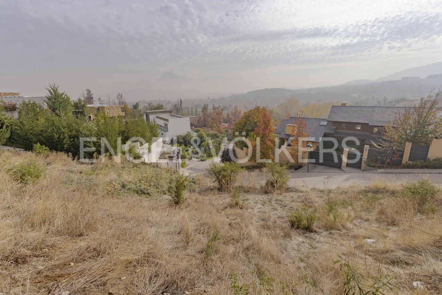 Hilly landscape with dry grass in the foreground, houses and trees in the midground, and mountains in the background under a cloudy sky.
