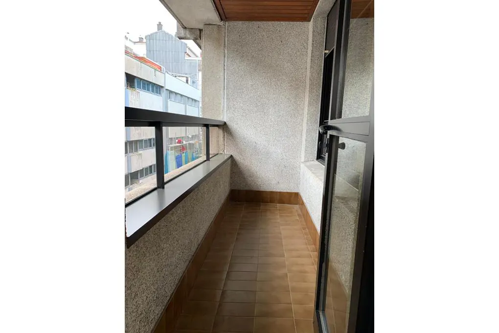A narrow balcony with brown tile flooring, a black metal railing, and a view of city buildings.