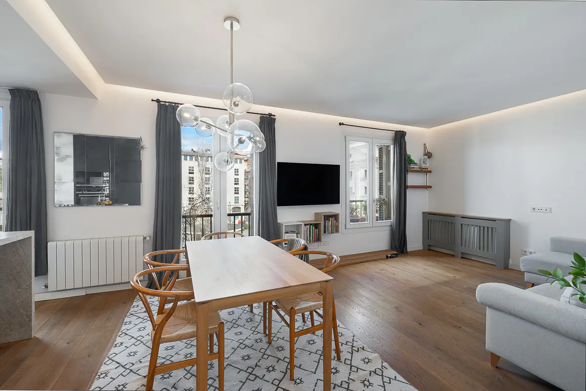 Bright living room with wood floors, a dining table with woven chairs, and a modern bubble chandelier. Gray curtains frame windows with city views.