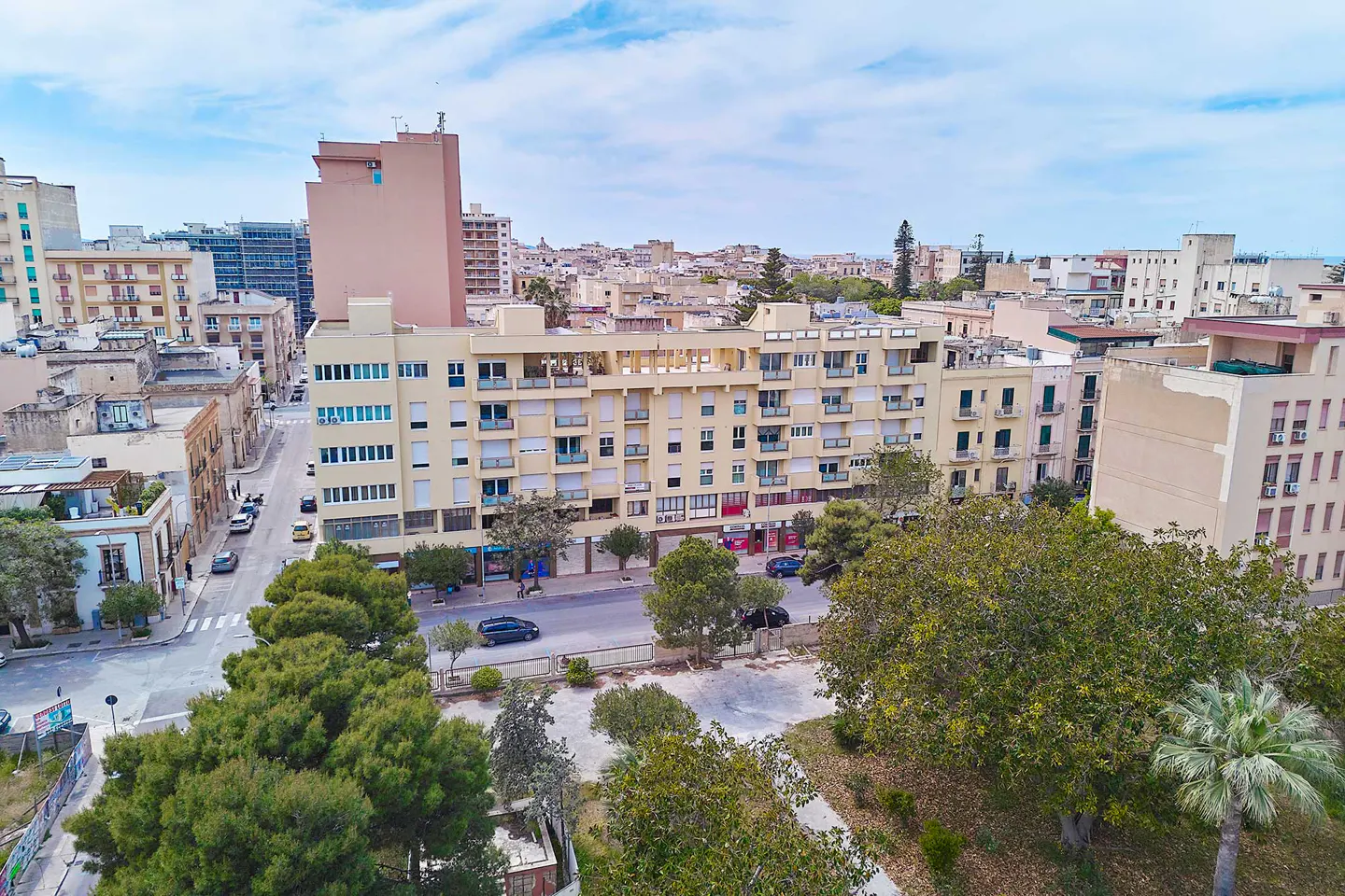 Aerial view of a city street with beige apartment buildings, green trees, and cars on the road under a blue sky.