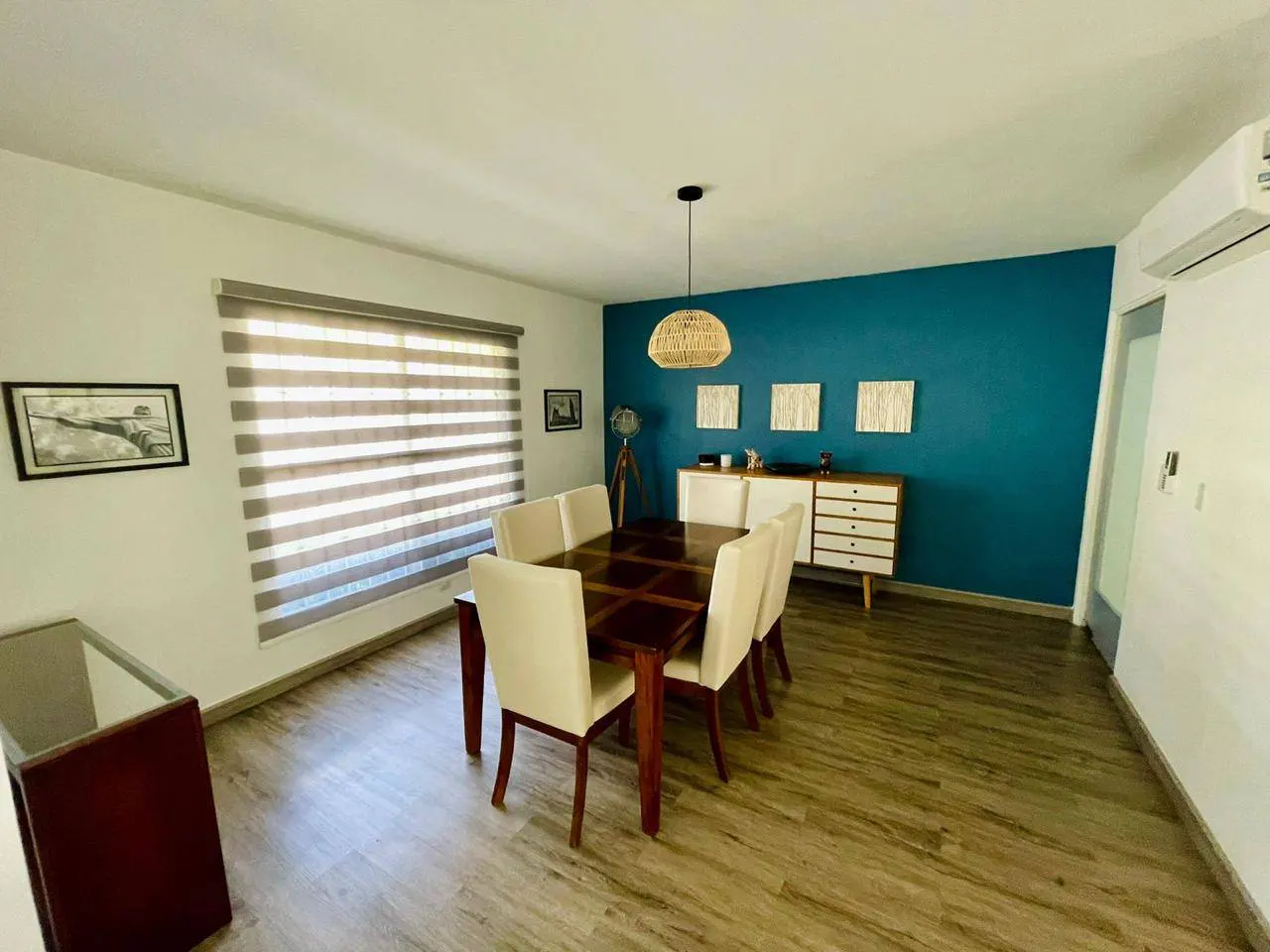 A dining room with a dark wood table, six cream chairs, and a woven pendant light. A blue accent wall and wood-look floors add warmth.