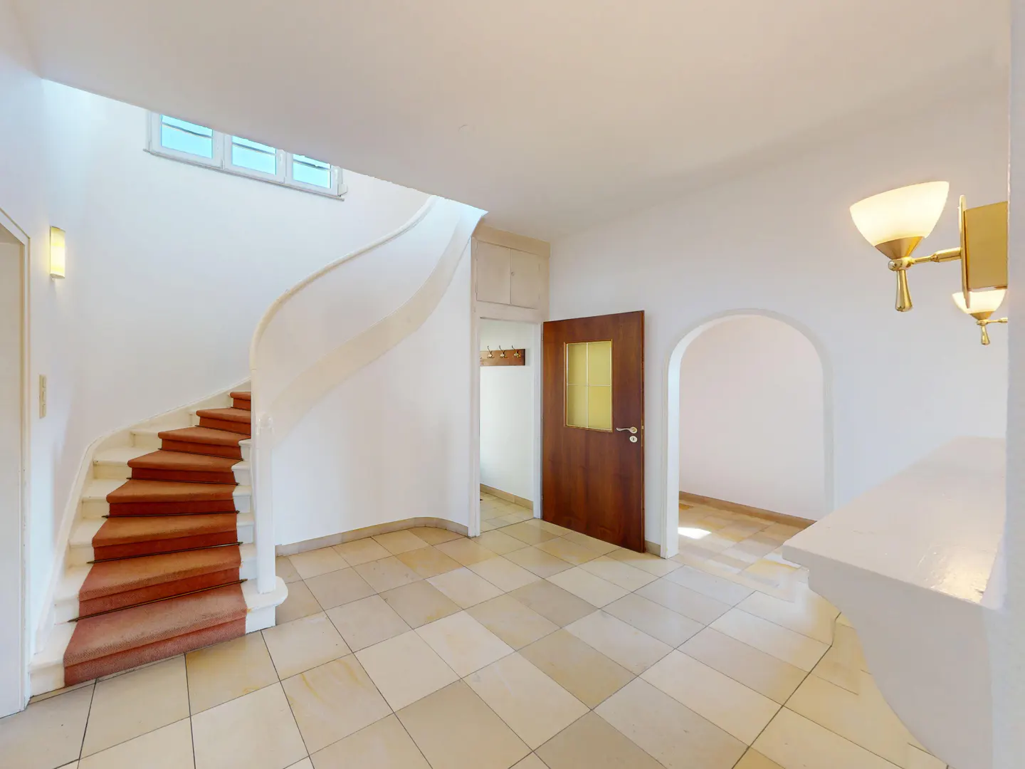 A bright foyer with a winding staircase, brown carpet, and tiled floor. A wooden door and arched doorway lead to other rooms.