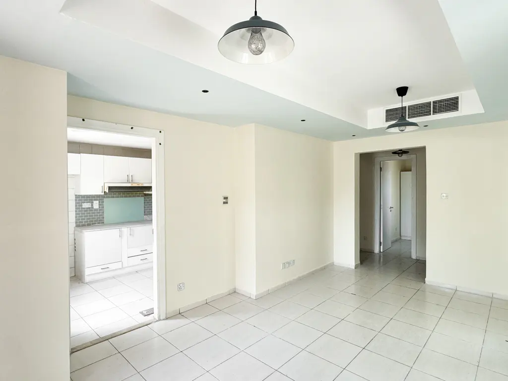 A bright, empty room with white walls and tile floors, lit by two pendant lights. A kitchen is visible through a doorway.