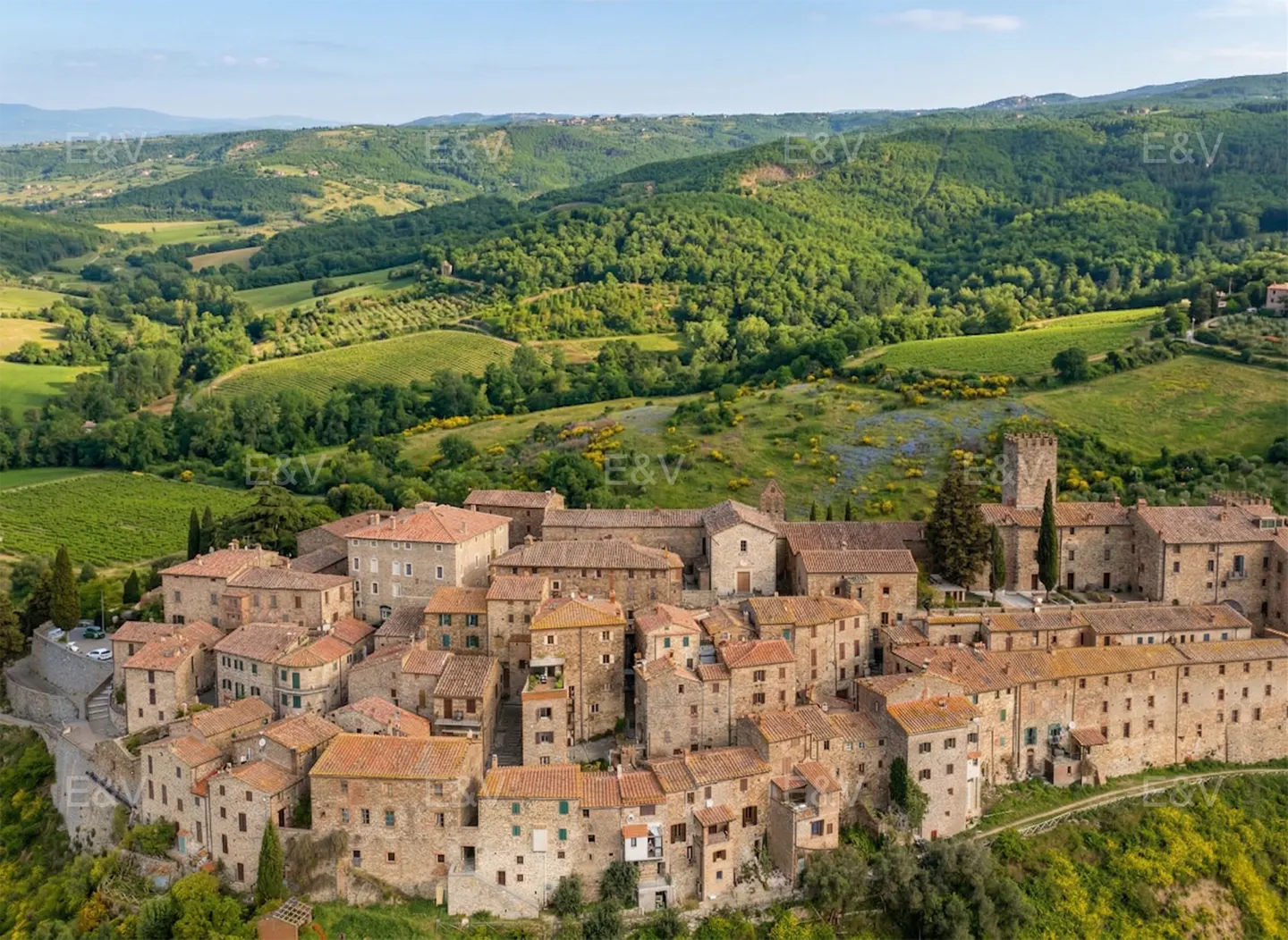 Aerial view of a Tuscan village with stone buildings and red tile roofs, nestled in green rolling hills.