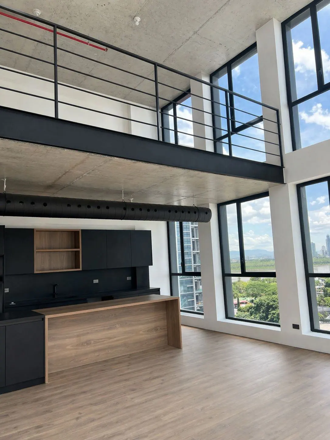 A modern apartment interior with black cabinets, a wood island, and large windows. A black metal railing overlooks the space.
