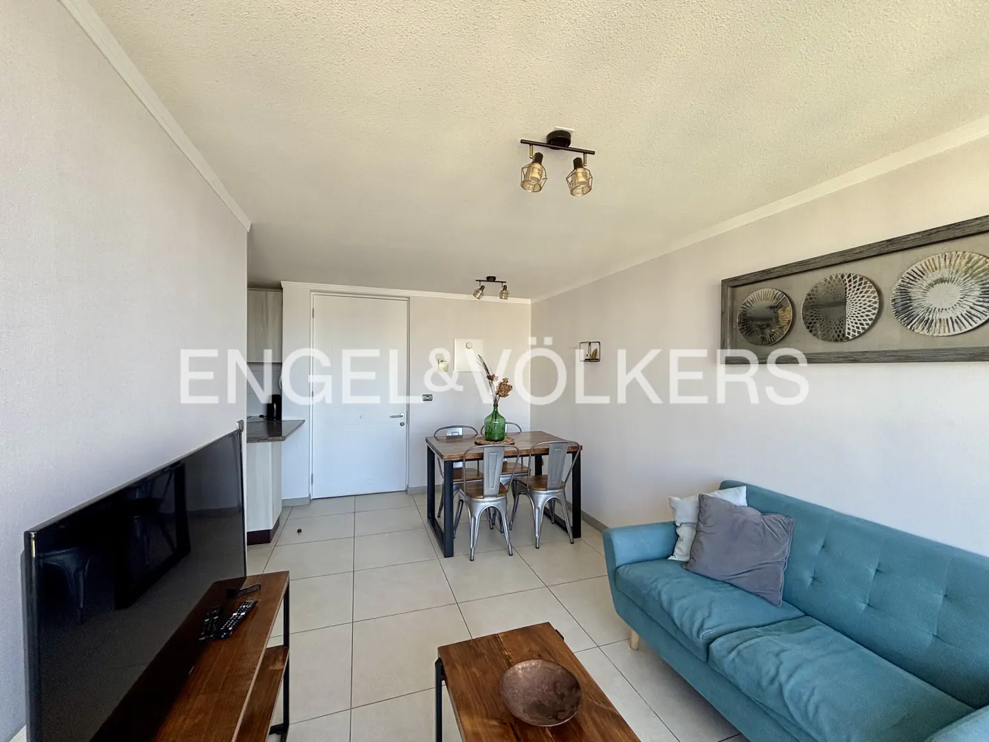Living room with a blue sofa, wooden table and chairs, and a TV. The walls are white, and there is a decorative picture on the wall.
